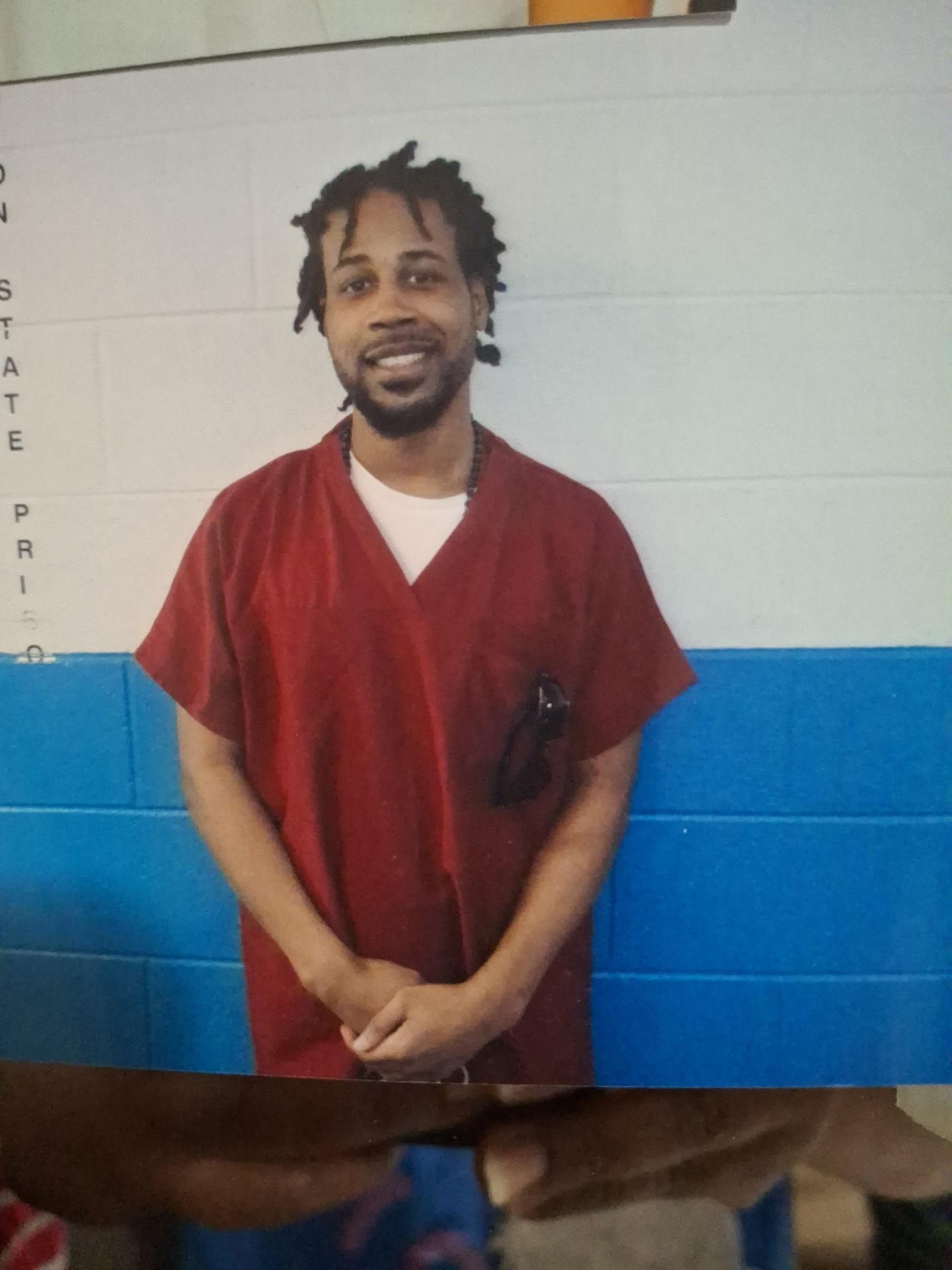 Dewaune Lane Jr. in a red prison uniform, smiling, standing against a white-and-blue wall inside a Virginia state prison