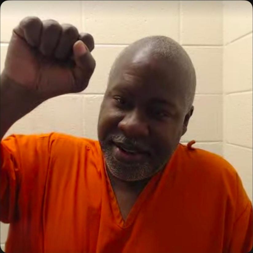 Kevin “Rashid” Johnson raises a clenched fist while wearing an orange prison uniform.